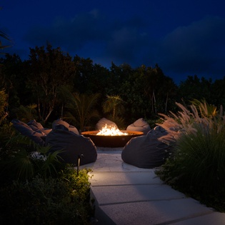 Evening gathers around a glowing fire pit surrounded by comfortable bean bag seating, framed by tropical plants—a perfect spot to unwind under the stars.