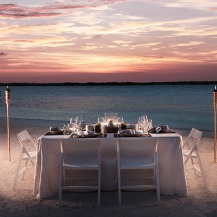 Romantic beachfront dining setup at sunset with elegant table, tiki torches, and stunning ocean views.