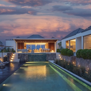 Modern villa features illuminated swimming pool and outdoor dining pavilion under dramatic twilight sky.