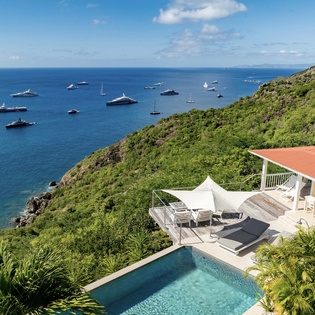 Stunning coastal villa perched above crystal-clear waters with luxury yachts dotting the horizon and lush tropical hillsides.