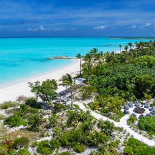 Aerial view of a pristine beachfront property showcasing turquoise waters, white sandy beaches, lush tropical vegetation, and serene outdoor lounging areas surrounded by palm trees and natural island beauty.