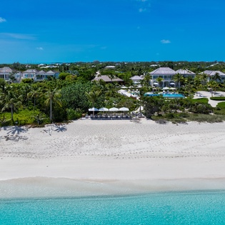 Aerial view of beachfront properties nestled among tropical foliage with white sand beach and turquoise waters beyond.