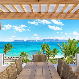 Shaded oceanfront dining terrace with turquoise sea views, tropical palms, distant mountain silhouettes, and comfortable wicker seating—perfect for relaxed meals in paradise.