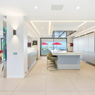 Your spacious modern kitchen flows seamlessly into the dining area, featuring sleek white cabinetry, premium appliances, and stunning water views through floor-to-ceiling windows.