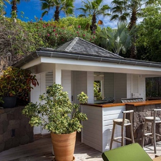 An outdoor bar pavilion nestled in a lush tropical garden, surrounded by palm trees and vibrant greenery under a brilliant blue sky.