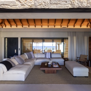 Sink into plush seating while exposed wooden beams frame your island retreat, with ocean views stretching beyond floor-to-ceiling windows.