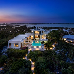 Aerial view of a modern waterfront estate at twilight, featuring a stunning pool, landscaped gardens, and expansive views over lagoons and natural surroundings.