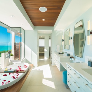 Soak in pure tranquility as sunlight streams through floor-to-ceiling windows, revealing breathtaking turquoise waters. Rose petals drift in your tub while warm wood tones and spa-like serenity surround you.