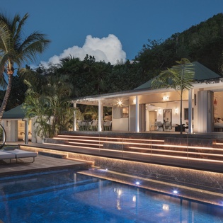 Stunning tropical villa with pool lighting creates magical evening atmosphere surrounded by palms and mountain views.