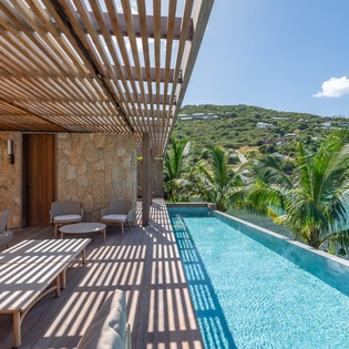Your private infinity pool overlooks turquoise Caribbean waters, with tropical palms swaying and shaded lounging beneath elegant wooden pergola.
