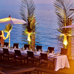 Elegant beachside dining area featuring illuminated palm trees and umbrellas, with ocean views and sunset ambiance creating a romantic tropical atmosphere.