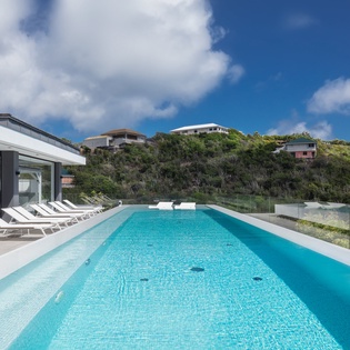 Modern villa featuring a pristine swimming pool surrounded by lush tropical hillside landscape under bright blue skies.