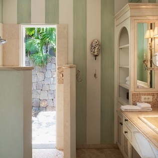 Step into your private sanctuary with elegant striped walls, a sunlit outdoor shower, and tropical garden views through the stone doorway.