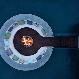 Stunning aerial view of a circular overwater fire pit with colorful seating, extending into pristine blue waters via wooden walkway.