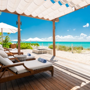 Beachfront deck features plush loungers with rolled towels under a flowing canopy, opening onto soft white sand and stunning turquoise waters—your perfect retreat for sun-soaked relaxation.