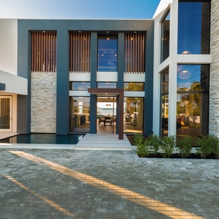 An impressive modern villa features striking architectural lines, textured stone walls, and warm lighting. The paved driveway leads to the welcoming entrance, flanked by tropical landscaping.