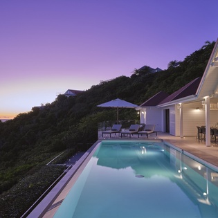 Hillside villa with infinity pool overlooking the ocean at sunset, surrounded by lush tropical vegetation and modern architecture.