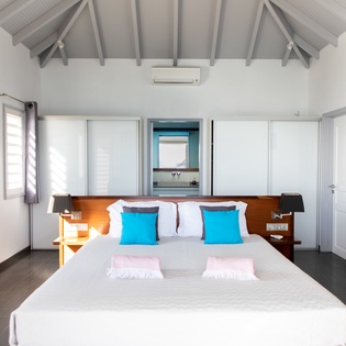 Unwind in your serene bedroom with soaring vaulted ceilings, crisp white linens, and vibrant blue accents that create a peaceful tropical retreat.