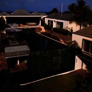 Modern tropical villa complex illuminated against twilight sky, showcasing contemporary architecture with traditional island roofing amid lush palm landscaping.