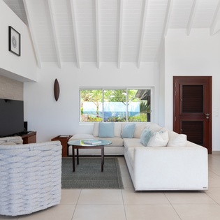 Sink into plush seating where soaring beamed ceilings and bright tropical light create your personal sanctuary. Watch sunlit palms sway through picture windows framing lush hillside views—your peaceful escape awaits.