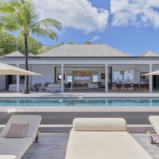 A spacious tropical villa featuring a pristine pool, sun-soaked loungers, and an open-air pavilion framed by swaying palms and lush greenery under bright blue skies.