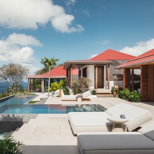 Luxurious beachfront villa with infinity pool overlooking the ocean, featuring red-tiled roofs and tropical landscaping.