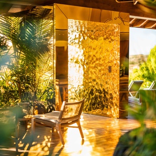 Sunlight filters through lush greenery onto your private terrace, where golden hour bathes the wooden deck and statement illuminated panel in warm, dreamy light.