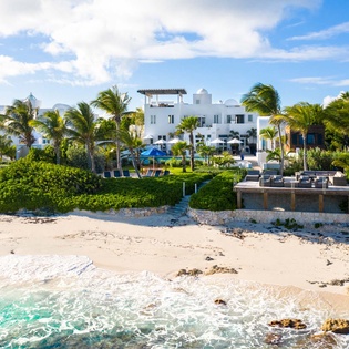 Stunning beachfront estate with white architecture, lush tropical gardens, and turquoise waters meeting sandy shores under bright blue skies.