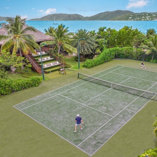 Aerial view of tennis court surrounded by tropical palm trees with ocean and mountain views in background.