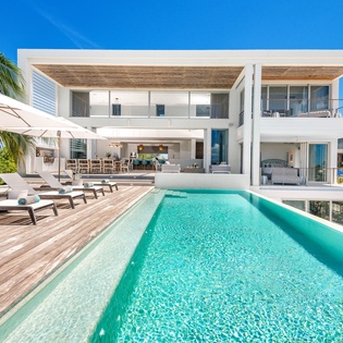 Modern white villa with infinity pool, wooden deck loungers, and tropical palms under brilliant blue Caribbean skies.