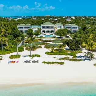 Beachfront estate nestled among lush tropical palms with turquoise waters, pristine sand, and a private pool surrounded by manicured gardens and coastal greenery.