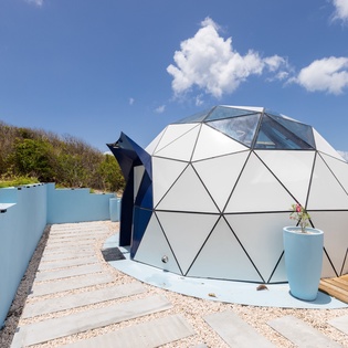 A striking geodesic dome perched on a clifftop deck overlooking the sparkling ocean, with sunny skies and modern outdoor spaces surrounding the unique structure.