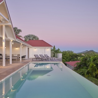 Modern villa with infinity pool overlooking tropical mountains at twilight, featuring covered terrace and elegant outdoor lounging areas.