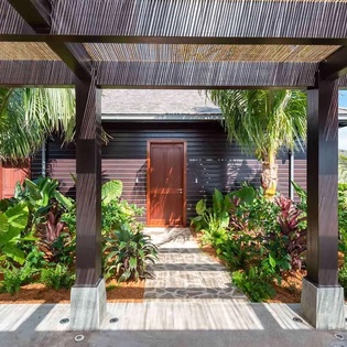 Tropical garden walkway leads to private entrance, framed by swaying palms and vibrant foliage under a natural bamboo pergola.