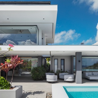 Modern villa features sleek architecture with tropical gardens and sparkling pool under brilliant blue skies.
