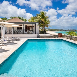 The beachfront property features a pristine swimming pool overlooking turquoise Caribbean waters, with elegant cabana lounging and tropical landscaping framing the coastal setting.