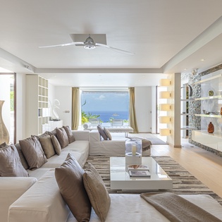 Relax in your elegant living room where bright coastal light and calming ocean blues greet you through floor-to-ceiling windows—your perfect spot to unwind.
