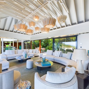 Your spacious living room features soaring exposed beam ceilings, floor-to-ceiling windows revealing lush tropical gardens, and plush seating perfect for relaxation.