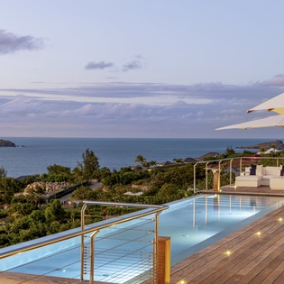 Stunning infinity pool overlooking lush tropical landscape and distant sea views at twilight, with elegant poolside lounge seating and umbrella shaded areas for relaxation.