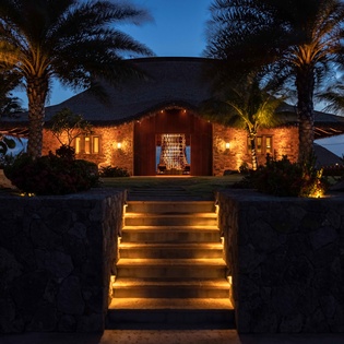 Stone villa with illuminated entrance stairs and tropical palms creates a stunning arrival under twilight skies.