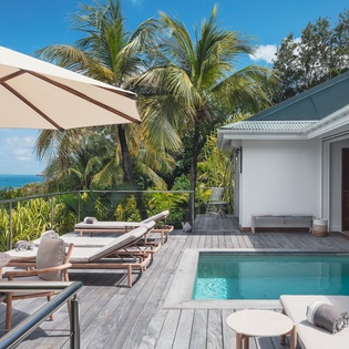 Expansive deck overlooking turquoise waters, with a private pool, sun loungers under shaded umbrella, and lush tropical palms framing your serene coastal retreat.