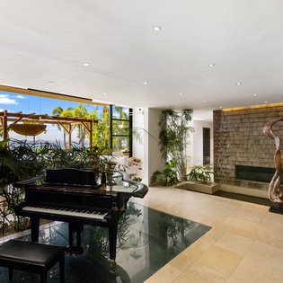 Your elegant living space features a grand piano, artistic fireplace, and floor-to-ceiling windows framing tropical views.