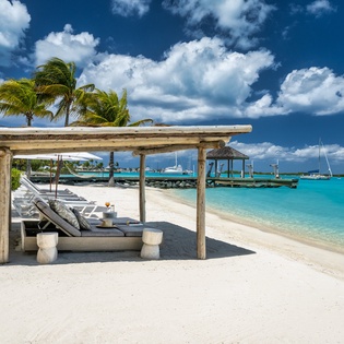 Private beachfront paradise with crystal-clear turquoise waters, white sand, and shaded loungers under swaying palms.