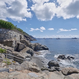 Rocky coastline with clear blue waters, stone pathways, and boats anchored offshore create a picturesque Mediterranean setting.