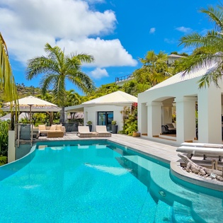 A contemporary tropical villa surrounded by lush hillside greenery, featuring an elegant curved pool flanked by swaying palms and pristine white architecture under brilliant blue skies.