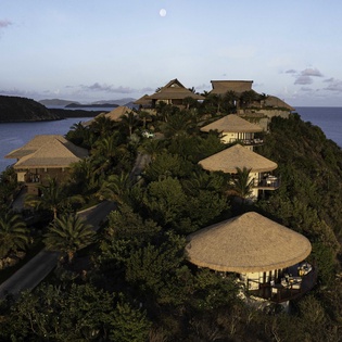 Tropical resort buildings perched dramatically on a lush hillside peninsula, surrounded by pristine blue waters and natural vegetation.