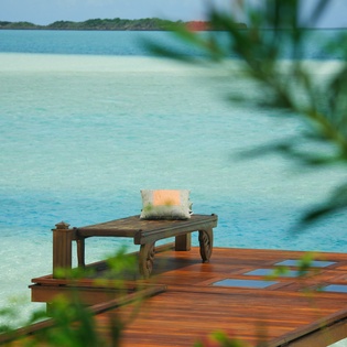 Your private deck extends over crystal-clear turquoise waters, where gentle waves lap beneath as you relax on the intimate wooden seating.