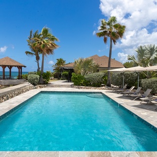 This beachfront villa features a pristine pool framed by swaying palms, with a gazebo overlooking turquoise waters and sandy shores just steps away—a tranquil Caribbean escape.