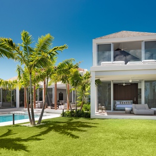 Modern beachfront villa with pristine lawn, swaying palms, and sparkling pool under brilliant blue skies.
