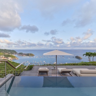 An infinity pool and lounge deck offering panoramic views of a coastal bay dotted with sailboats and lush hillsides. Perfect for relaxation and taking in breathtaking sunsets.